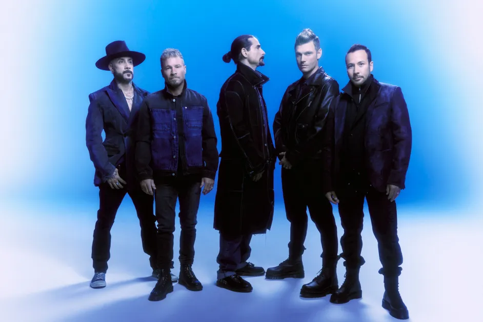 The five members of Backstreet Boys standing together in dark outfits against a blue backdrop