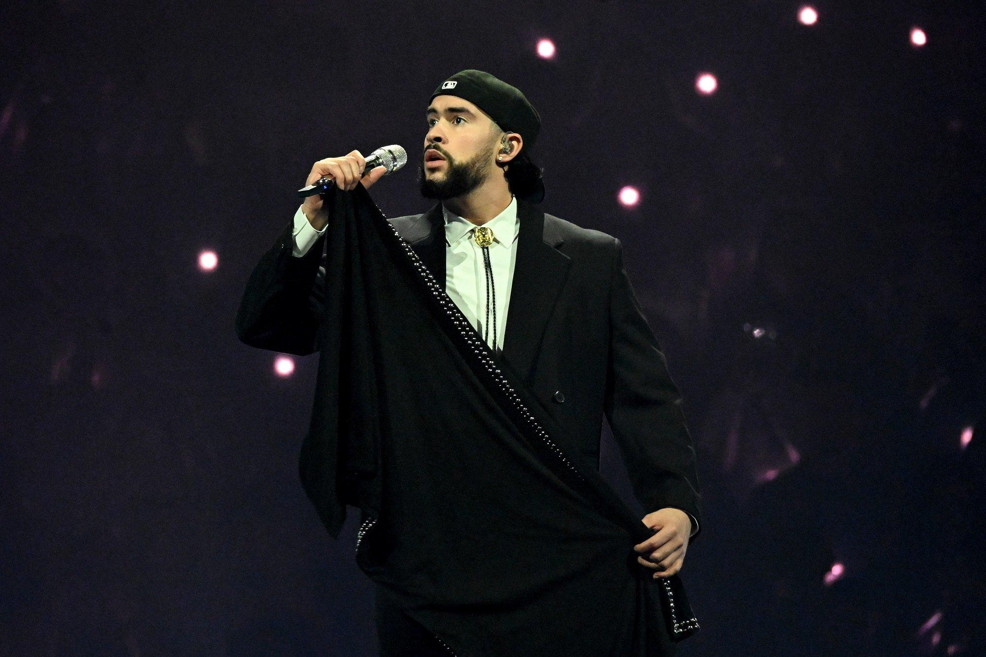 Bad Bunny singing into a microphone on stage in a dark suit and bolo tie
