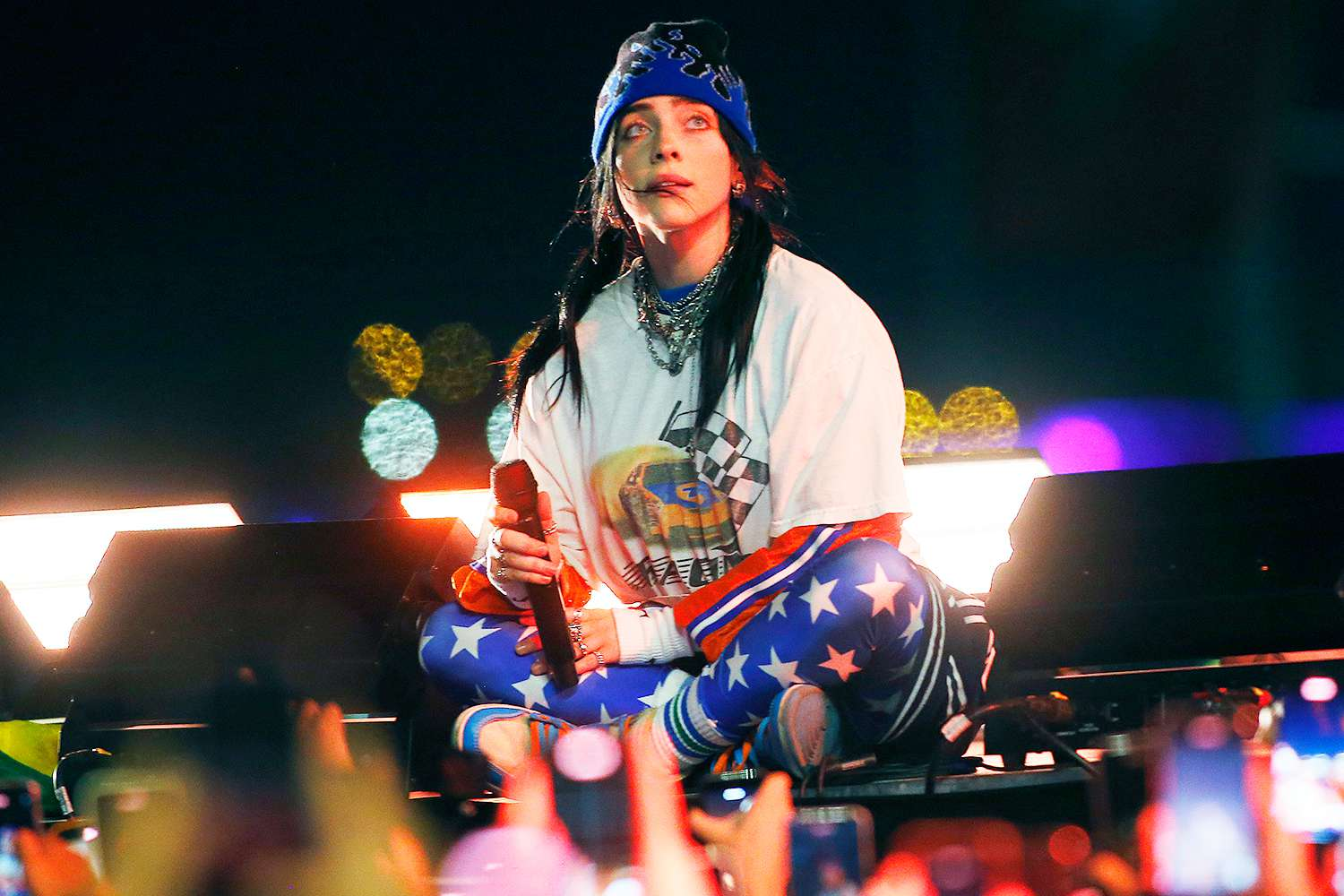 Billie Eilish performing live, seated, holding a microphone in front of a crowd