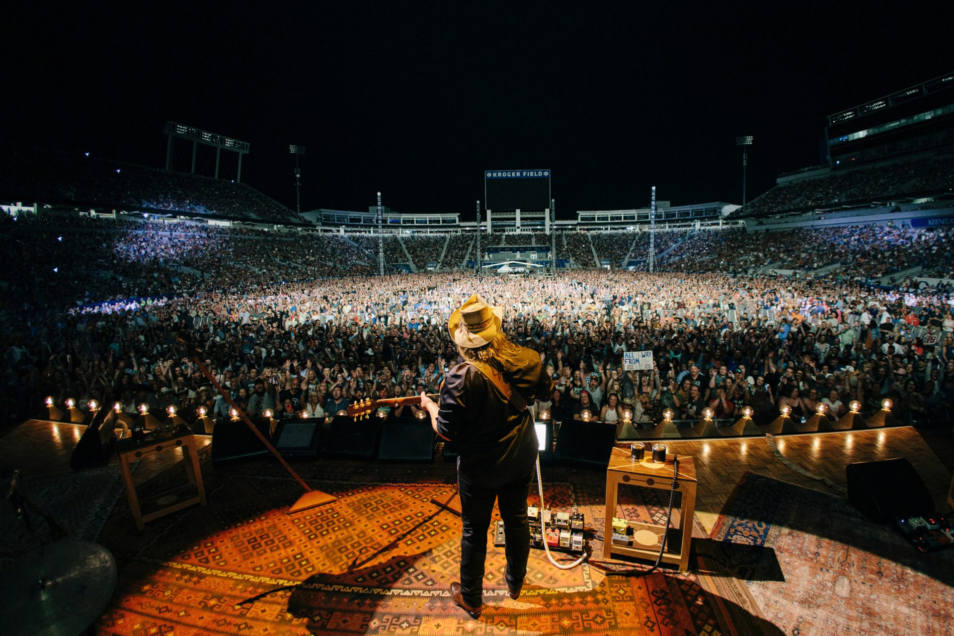 A performer viewed from behind on stage facing a packed football stadium crowd at night