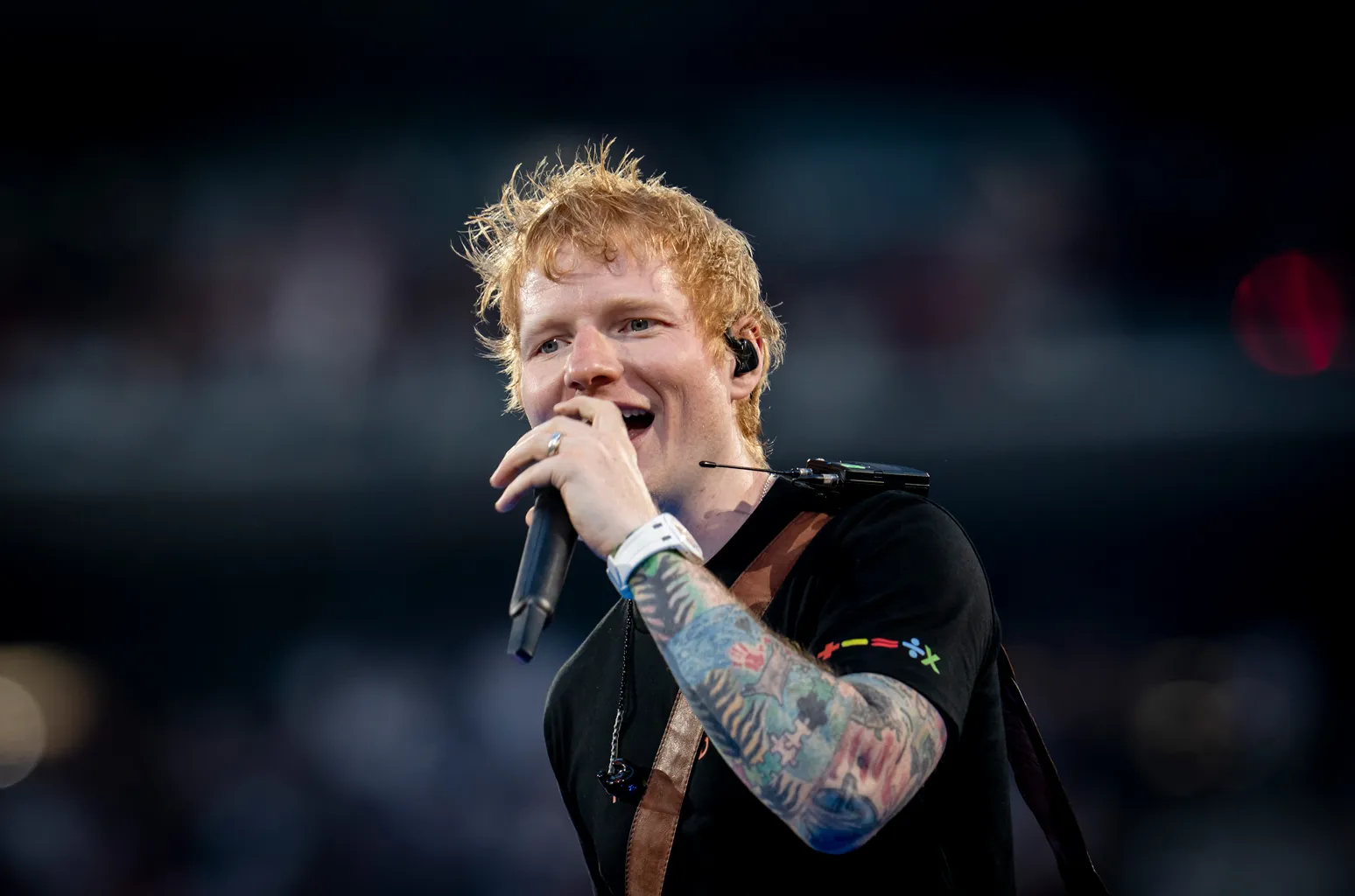 Ed Sheeran singing into a microphone on stage, tattooed arm raised, with a stadium crowd blurred behind him