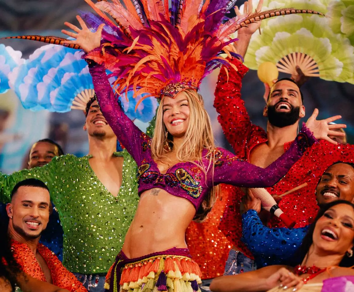 Karol G on stage in a feathered carnival-style headdress and costume, surrounded by dancers in colourful sequined outfits