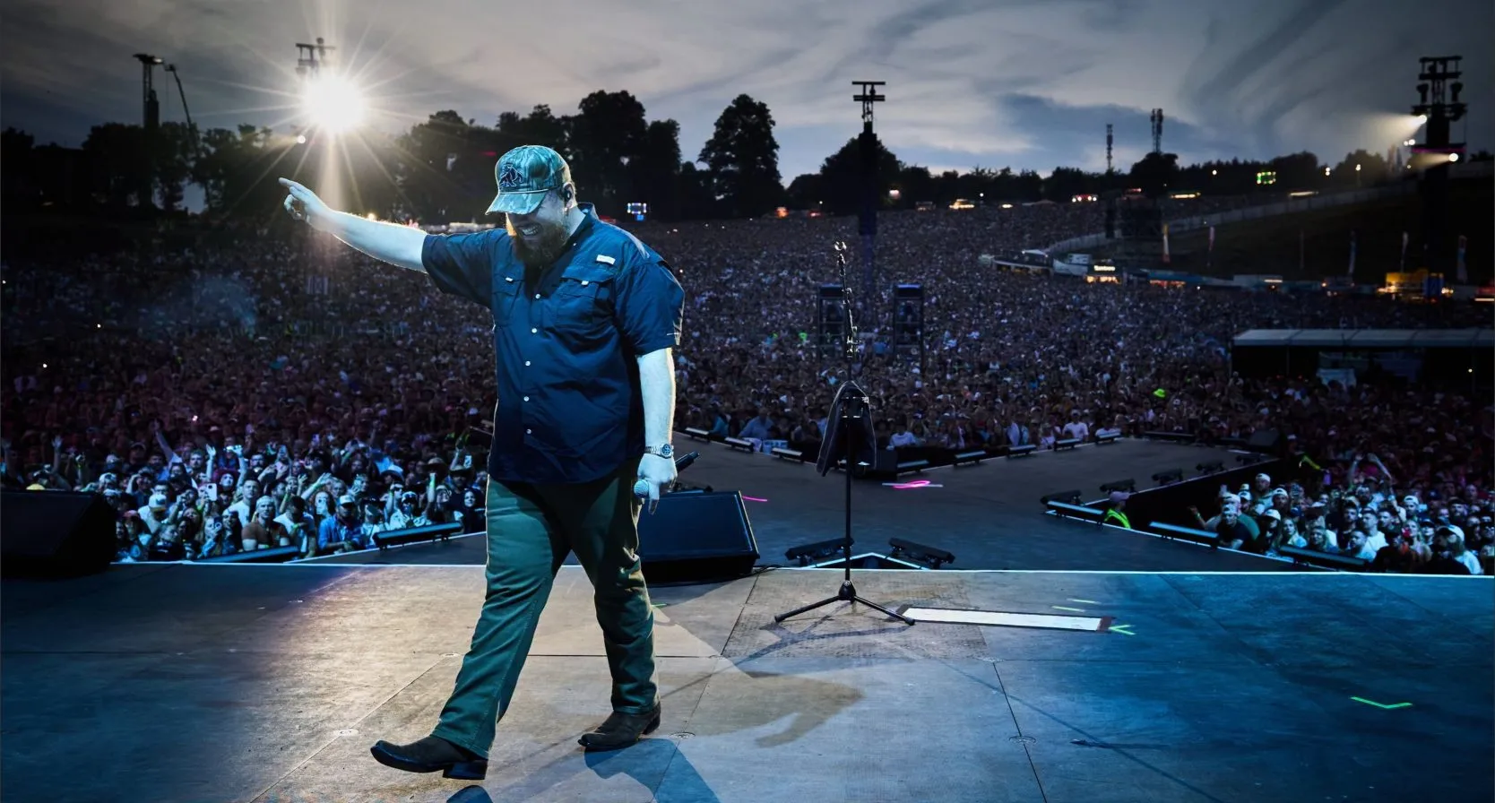 Luke Combs performing on an outdoor stage at dusk, pointing to a packed stadium crowd