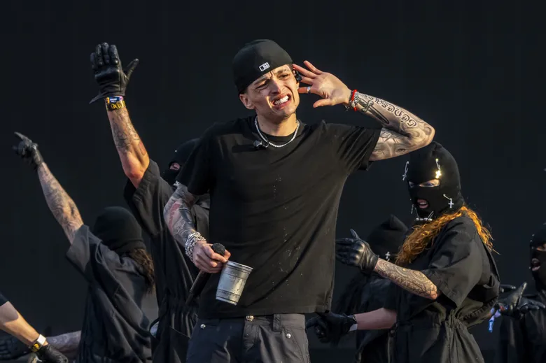A performer on stage in a black beanie and t-shirt, arm raised, with masked backup performers behind him