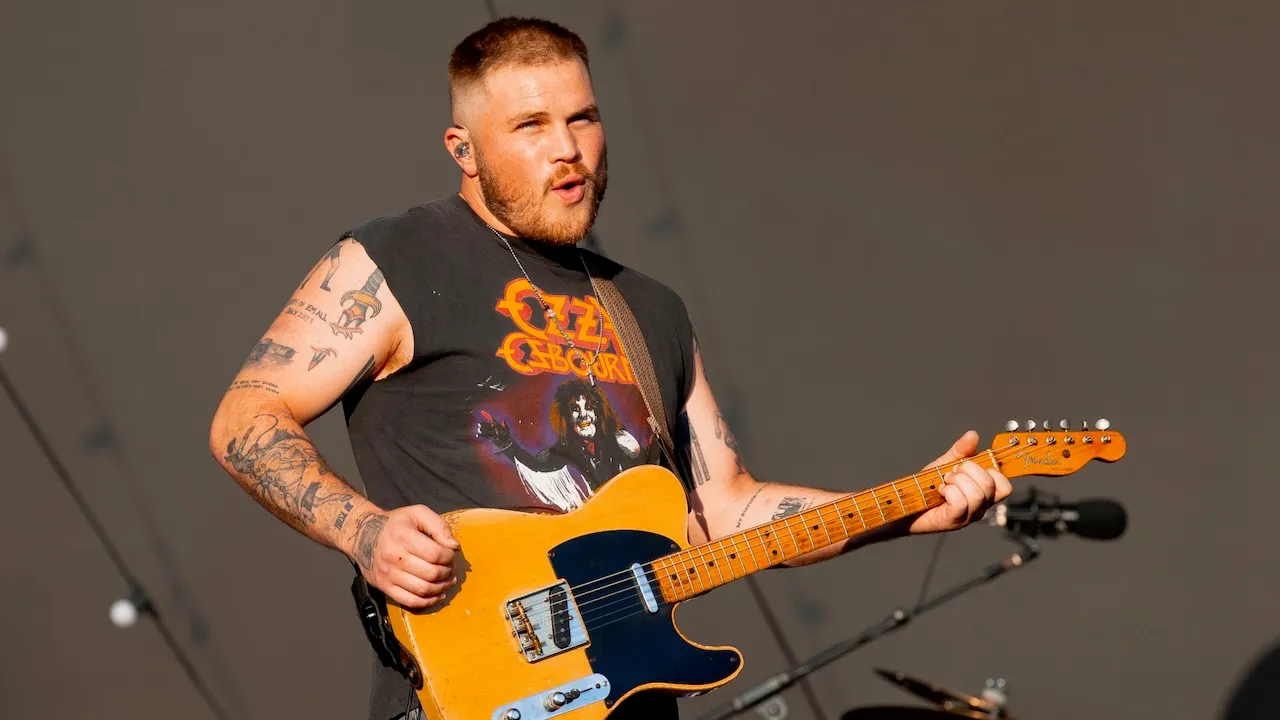 Zach Bryan viewed from behind on stage, playing guitar facing a packed stadium crowd at night